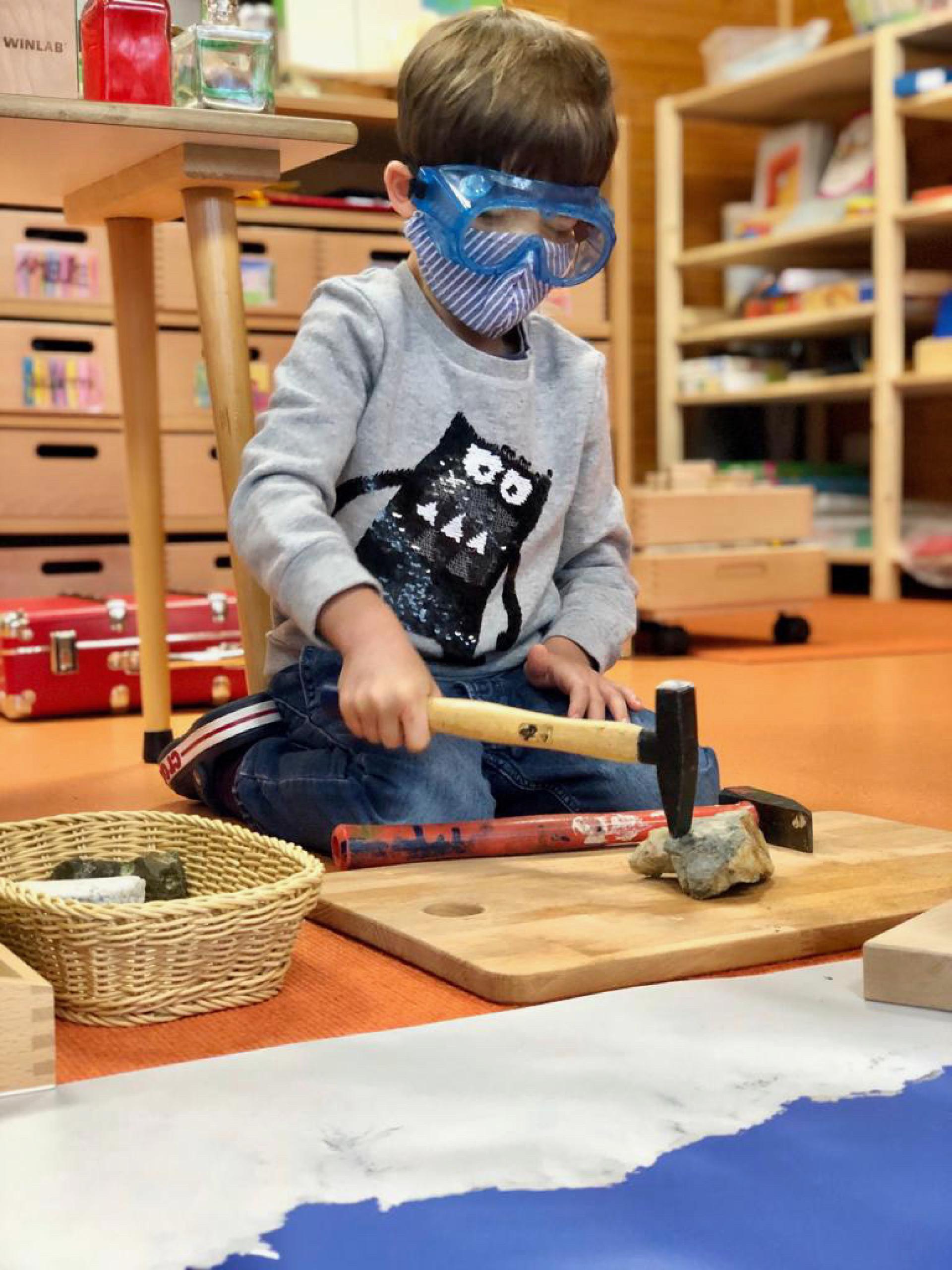 Ein Junge in Maske und Brille bearbeitet einen Stein mit einem Hammer