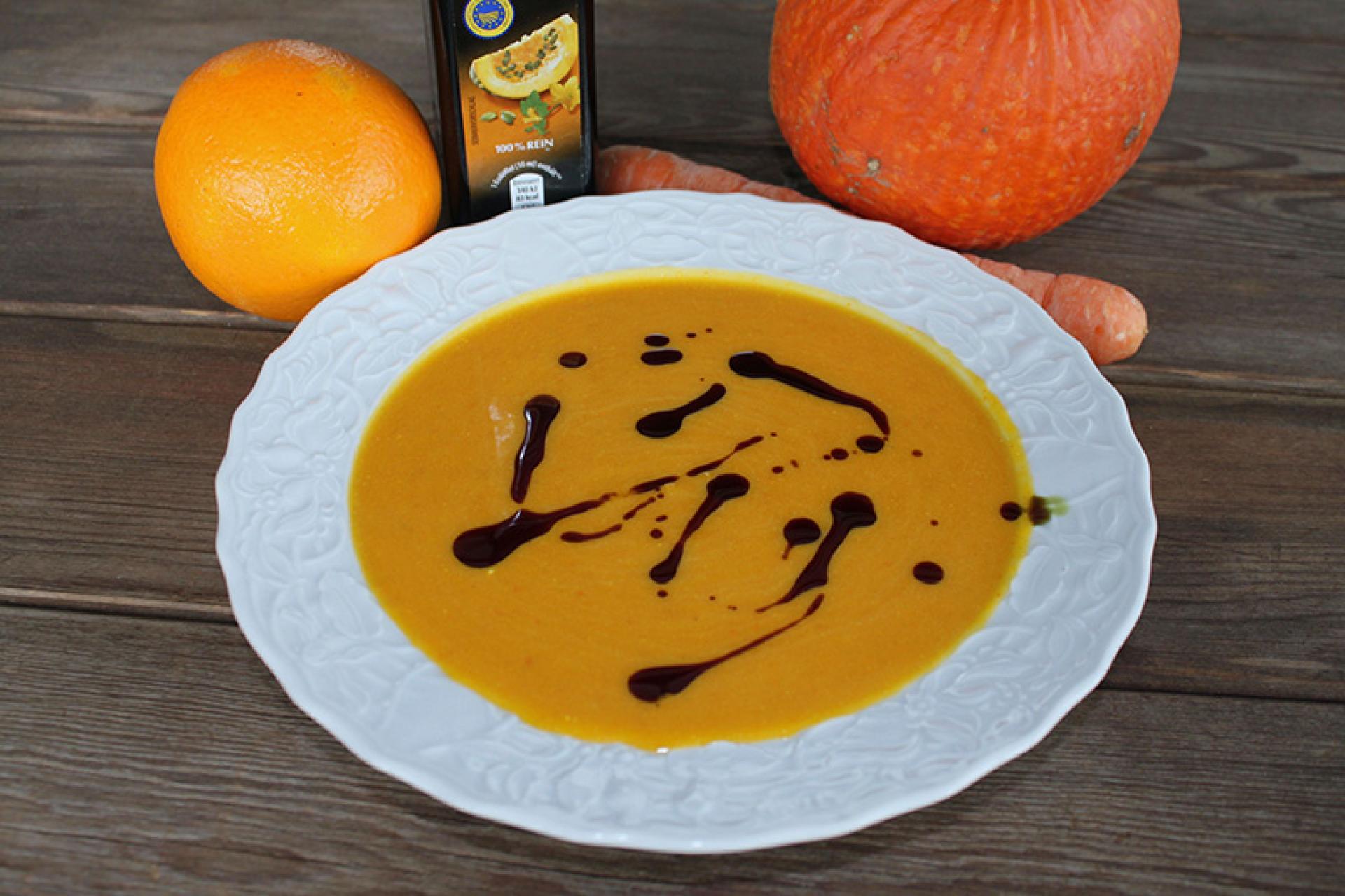 Eine Teller Kürbissuppe, eine Orange, eine Möhre und ein Kürbis auf einem Tisch arrangiert