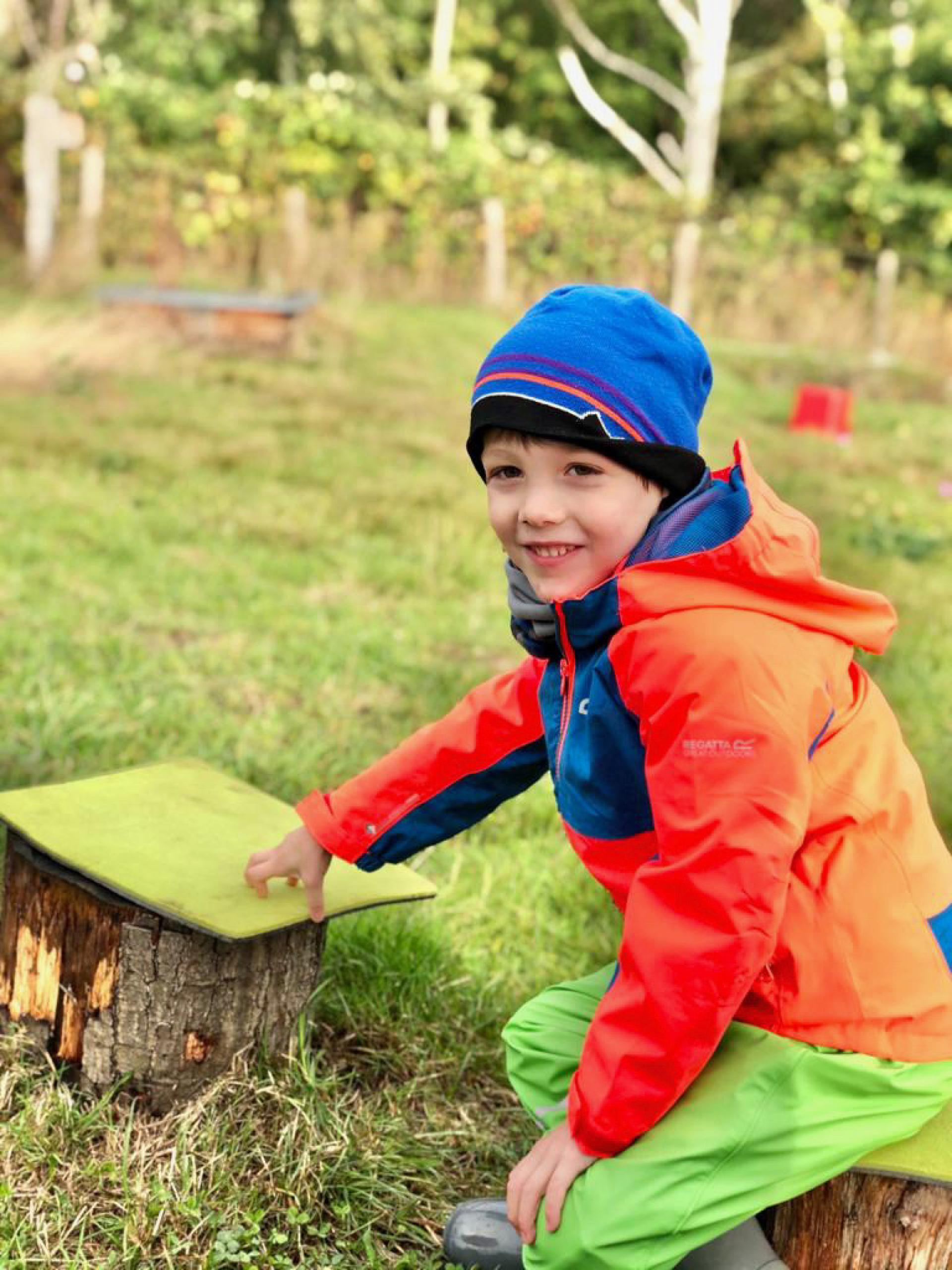 Ein Junge in einer orangefarbenen Jacke und blauen Mütze sitzt auf einem Baumstamm